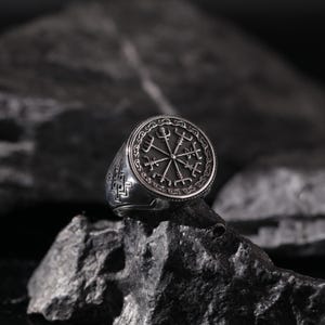 May include: A silver ring with a black compass design featuring a circle of runes. The ring is on a dark gray rock.