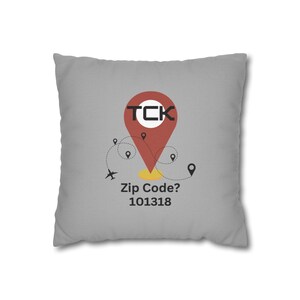 Puede incluir: Un cojín cuadrado gris con un gráfico de chincheta roja. La chincheta tiene las letras "TCK" dentro de un círculo blanco. Debajo de la chincheta está el texto "Zip Code? 101318". Hay pequeñas chinchetas negras y una línea punteada con un avión.