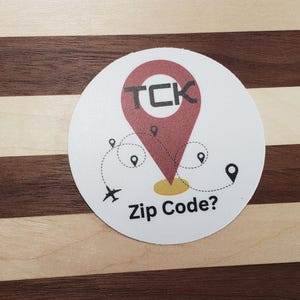 Puede incluir: Una pegatina redonda blanca con un gráfico de chincheta roja y el texto "TCK" en negro. La pegatina también presenta el texto "Zip Code?" y una línea punteada con pequeños iconos de chincheta y un avión.