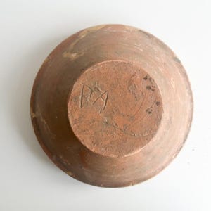 Pu&ograve; includere: Una ciotola in ceramica fatta a mano, color terracotta, vista dal basso. La ciotola ha una forma ampia e poco profonda con una superficie leggermente strutturata. La base presenta un disegno inciso, forse iniziali o un marchio del produttore.