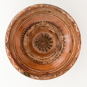 Könnte beinhalten: Eine runde Keramikschale in Terrakotta-Farbe mit detailliertem Design. Die Schale weist konzentrische Kreise und ein zentrales sonnenähnliches Motiv auf. Weiße und braune Akzente heben den Rand und die Innenmuster hervor. Die Schale hat ein rustikales, handgefertigtes Aussehen.
