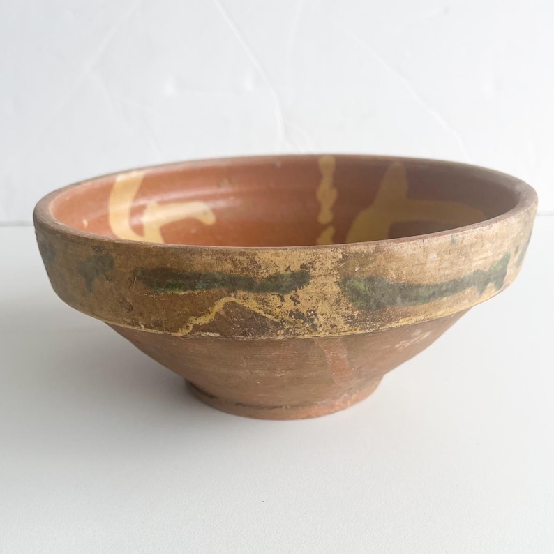 Pu&ograve; includere: Una ciotola rustica in terracotta con un bordo largo e un corpo dolcemente inclinato. La ciotola presenta un colore marrone caldo e terroso con accenti decorativi gialli e verdi attorno al bordo. Il diametro della ciotola &egrave; di circa 25 cm.