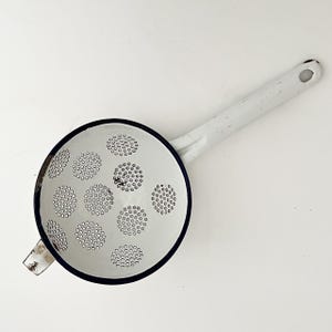May include: A white enamelware sieve with a long handle and a dark blue rim. The sieve features many small holes for draining. The handle has a hole for hanging. A classic kitchen utensil.