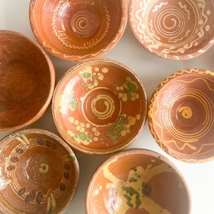 Pode incluir: Uma coleção de tigelas de terracota feitas à mão com vários designs. As tigelas são principalmente laranja-castanho com detalhes brancos e verdes. Algumas tigelas apresentam padrões em espiral e circulares, enquanto outras têm motivos florais ou geométricos. As tigelas variam em tamanho e forma.