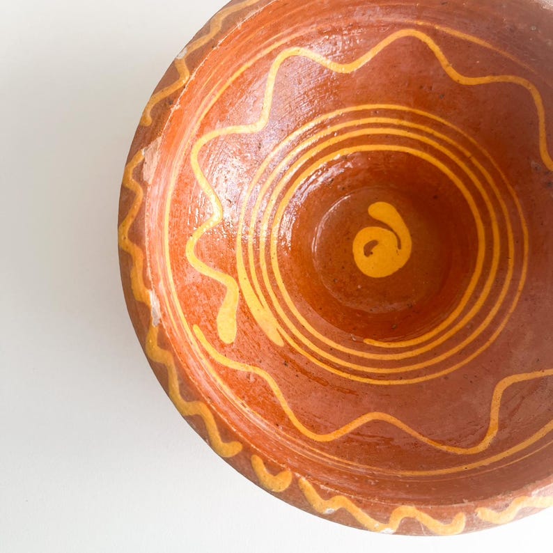 Pu&ograve; includere: Una ciotola in terracotta fatta a mano con una base marrone caldo e disegni a spirale e ondulati dipinti di giallo. L'interno della ciotola presenta cerchi concentrici e un vortice centrale, mostrando un'estetica rustica e artigianale.