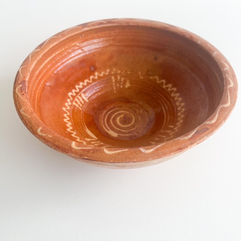 Pu&ograve; includere: Una ciotola rustica color terracotta con un bordo largo e un interno poco profondo. La ciotola presenta un motivo decorativo a zig-zag bianco sul bordo e un disegno a spirale al centro. La ciotola &egrave; probabilmente fatta a mano e ha un'estetica naturale e terrosa.