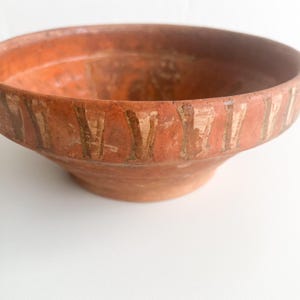 Pu&ograve; includere: Una ciotola in terracotta con un design ampio e poco profondo. La ciotola ha un colore marrone-rossastro con segni verticali sbiaditi e di colore chiaro attorno al bordo. La ciotola &egrave; su una superficie bianca.