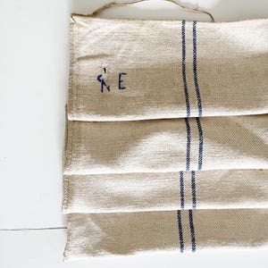 Puede incluir: Cuatro toallas de té de lino dobladas con rayas azules y las iniciales bordadas "K.E."