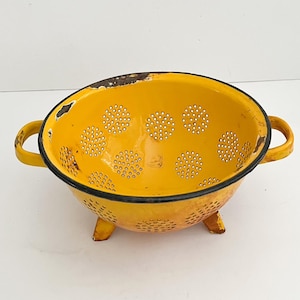May include: A vintage yellow colander with a black rim and two handles. The colander has small holes in a circular pattern and three small feet. The enamel is chipped in places, giving it a rustic appearance.