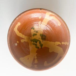 Pu&ograve; includere: Una ciotola in ceramica fatta a mano con esterno e interno marrone rossiccio. L'interno presenta un design astratto giallo chiaro con accenti verdi. Il bordo della ciotola &egrave; di una tonalit&agrave; di marrone pi&ugrave; scura e l'estetica generale suggerisce uno stile rustico e artigianale.