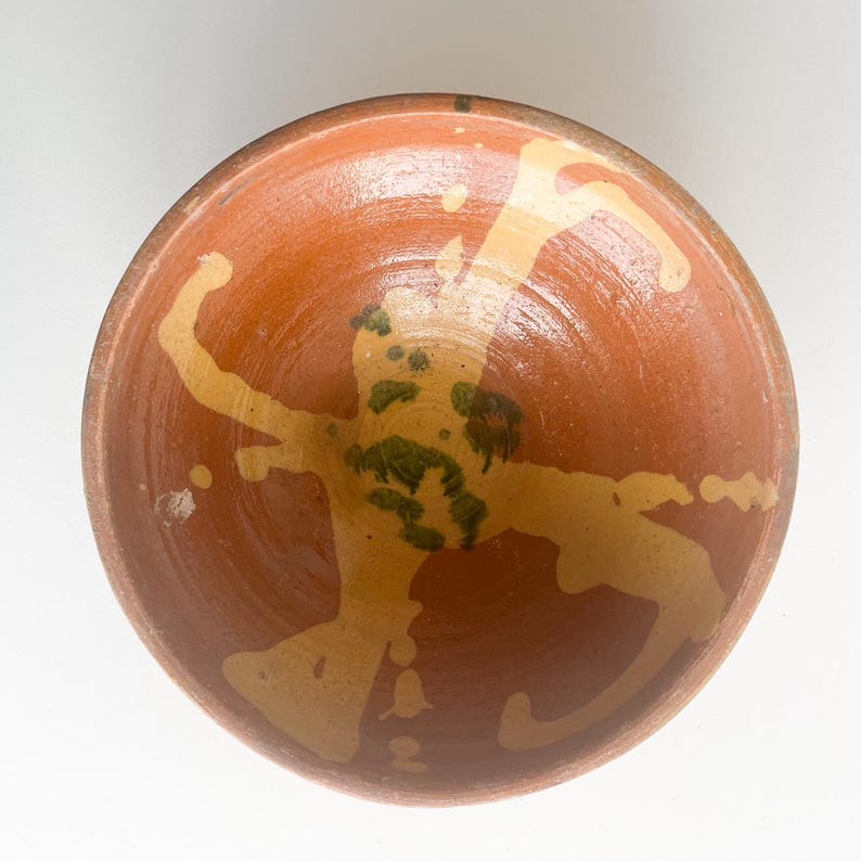 Pu&ograve; includere: Una ciotola in ceramica fatta a mano con esterno e interno marrone rossiccio. L'interno presenta un design astratto giallo chiaro con accenti verdi. Il bordo della ciotola &egrave; di una tonalit&agrave; di marrone pi&ugrave; scura e l'estetica generale suggerisce uno stile rustico e artigianale.
