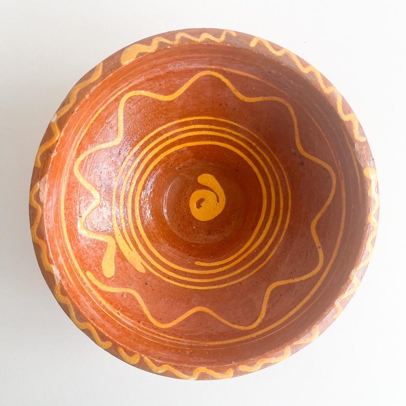 Pu&ograve; includere: Una ciotola fatta a mano color terracotta con disegni dipinti di giallo. La ciotola presenta un motivo ondulato sul bordo e cerchi concentrici con una spirale al centro. L'interno &egrave; di una tonalit&agrave; di marrone pi&ugrave; scura.