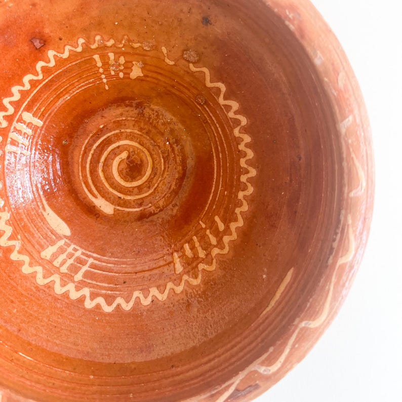 Pu&ograve; includere: Primo piano di una ciotola color terracotta con un disegno bianco a spirale e ondulato al centro. L'interno della ciotola presenta cerchi concentrici e una finitura lucida. Il bordo ha un motivo bianco ripetuto.