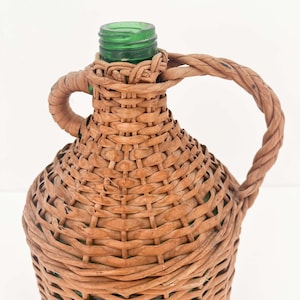 May include: A green glass bottle encased in a woven light brown wicker covering. The bottle features a narrow neck with a threaded top and a single twisted wicker handle attached to the side and the neck of the bottle.