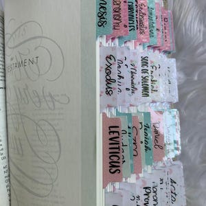 May include: A close-up of a Bible with colorful bookmarks for each book of the Old Testament. The bookmarks are decorated with different patterns and designs. The bookmarks are labeled with the names of the books of the Bible, including Genesis, Exodus, Leviticus, Numbers, Deuteronomy, Joshua, Judges, Ruth, 1 Samuel, 2 Samuel, 1 Kings, 2 Kings, 1 Chronicles, 2 Chronicles, Ezra, Nehemiah, Esther, Job, Psalms, Proverbs, Ecclesiastes, Song of Solomon, Isaiah, Jeremiah, Lamentations, Ezekiel, Daniel, Hosea, Joel, Amos, Obadiah, Jonah, Micah, Nahum, Habakkuk, Zephaniah, Haggai, Zechariah, Malachi.