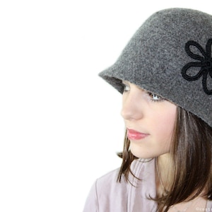 May include: A gray felt cloche hat with a black flower design. The hat has a wide brim and a rounded crown.