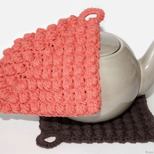 May include: A coral crocheted pot holder with a brown crocheted pot holder underneath. The coral pot holder is draped over a gray teapot.