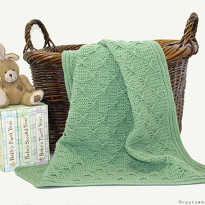 Puede incluir: Una manta de bebé de crochet verde con un patrón de estrellas está doblada y colocada en una cesta de mimbre. Tres libros titulados "Baby's First Year", "Baby's Second Year" y "Baby's Third Year" están apilados en una mesa junto a un conejito de peluche marrón.