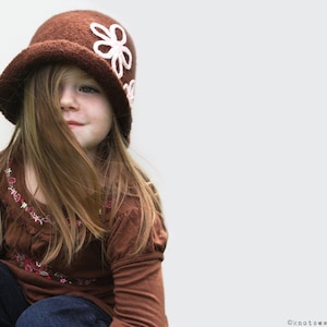 May include: A young person wearing a brown felt hat with white flower embellishments. The hat has a wide brim and is tilted slightly to the side. The person is wearing a brown long-sleeved shirt and blue jeans.