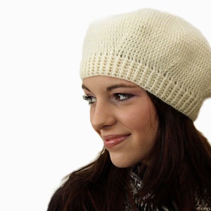 May include: A woman wearing a white crocheted beret. She is looking to the right and smiling.