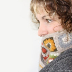 May include: A woman with brown hair wearing a gray sweater with a colorful crocheted scarf. The scarf has a geometric pattern with orange, yellow, green, and red accents.