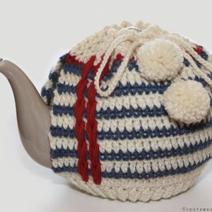 May include: A white, blue, and red striped crocheted teapot cozy with white pom-poms. The cozy is on a gray teapot.