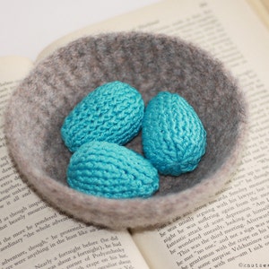 May include: Three teal blue crocheted eggs in a gray felt bowl. The bowl is sitting on top of an open book.