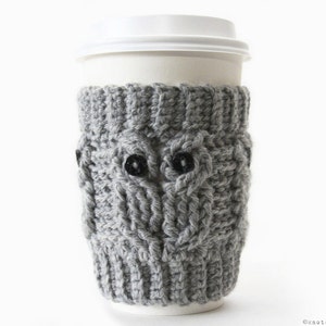 May include: Gray knitted coffee cup cozy with a cable knit pattern and two black buttons for eyes. The cozy is on a white paper coffee cup.
