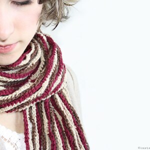 May include: A close-up of a woman wearing a striped scarf in shades of brown, beige, and burgundy. The scarf is crocheted and has a loose, flowing texture.