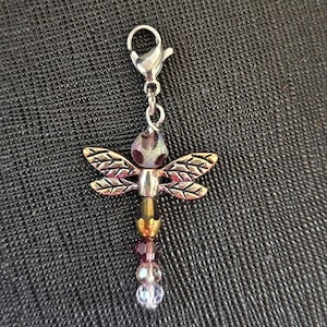 May include: A dragonfly charm with a silver clasp. The dragonfly has silver wings, a purple and iridescent body, and a beaded tail with purple, gold, and clear beads. The charm is designed to be attached to a bracelet or necklace.