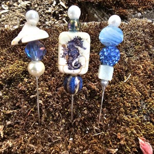 May include: Three decorative pins with blue and white beads and a seahorse design. The pins are on a bed of brown moss.