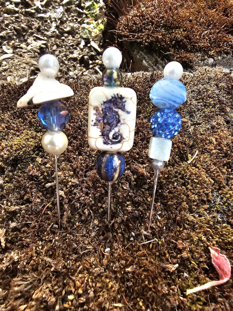 May include: Three decorative pins with blue and white beads and a seahorse design. The pins are on a bed of brown moss.