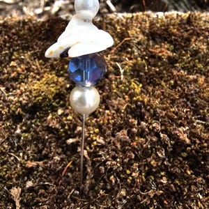 May include: A white ceramic figurine with a blue glass bead and a white pearl on a silver pin. The pin is stuck into a bed of brown moss.
