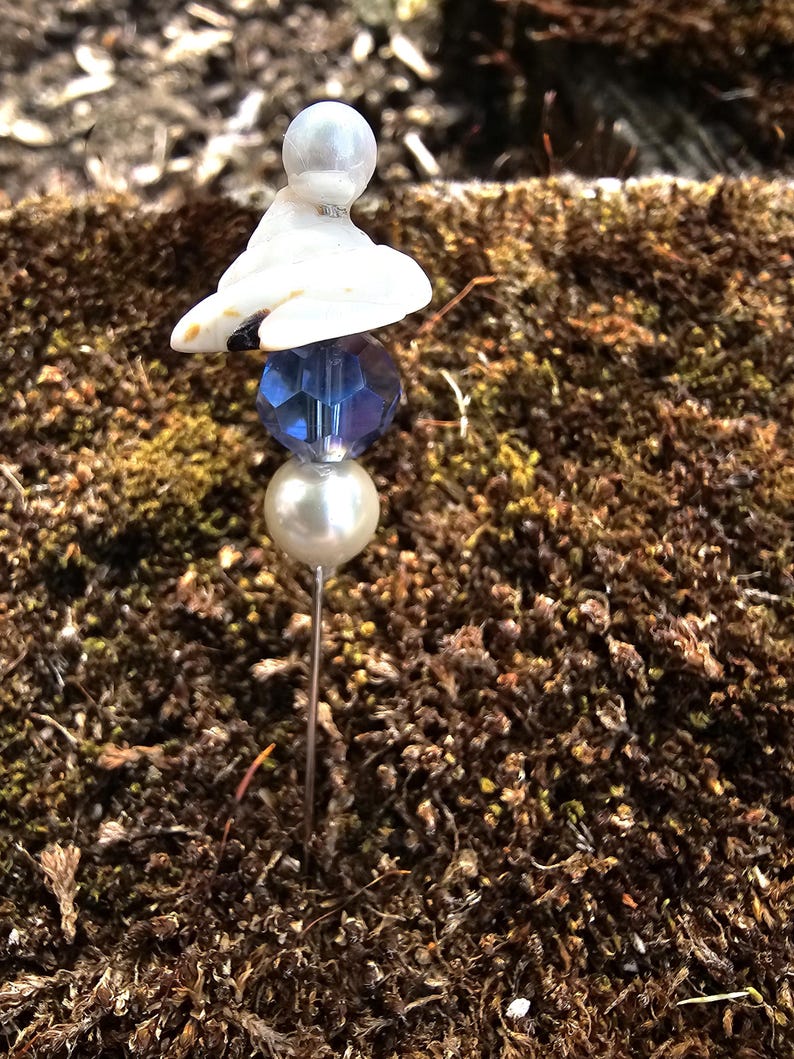 May include: A white ceramic figurine with a blue glass bead and a white pearl on a silver pin. The pin is stuck into a bed of brown moss.