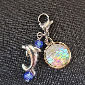 May include: A silver-colored dolphin charm with blue crystal beads and a round charm with iridescent scales. The charm is attached to a silver-colored clasp, suitable for a bracelet or necklace.