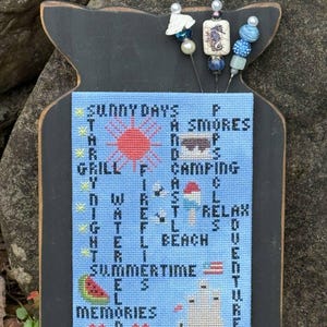 May include: A blue and white cross-stitch design on a black wooden plaque with the words "Sunny Days", "Grill", "Water", "Summertime", and "Memories" incorporated into the design. The design also includes a red sun, a campfire, a beach umbrella, a watermelon, and a sandcastle.