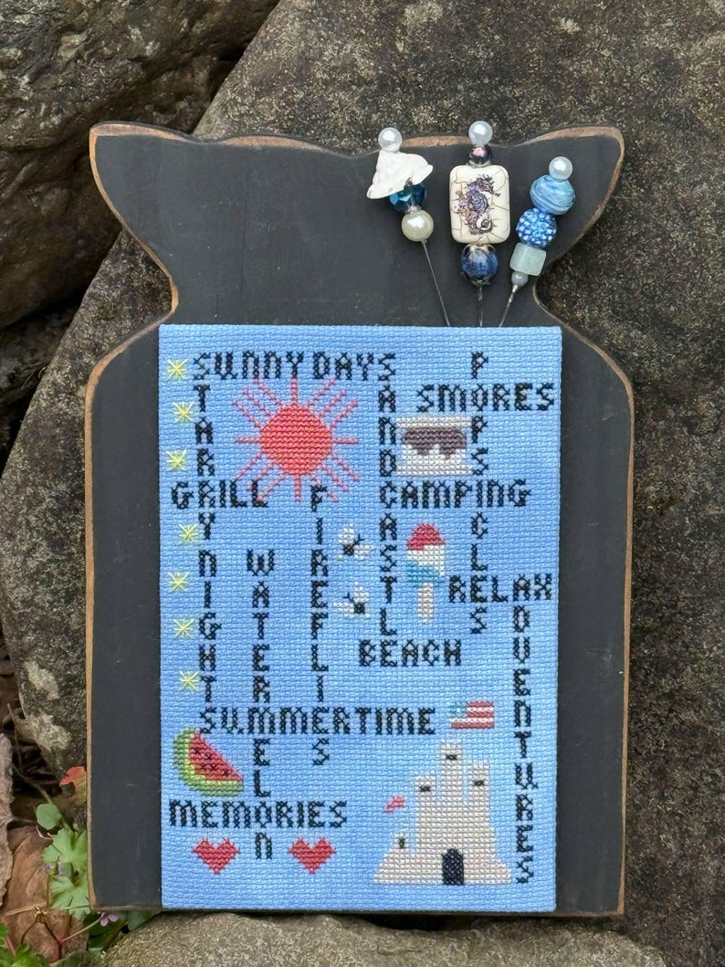 May include: A blue and white cross-stitch design on a black wooden plaque with the words "Sunny Days", "Grill", "Water", "Summertime", and "Memories" incorporated into the design. The design also includes a red sun, a campfire, a beach umbrella, a watermelon, and a sandcastle.
