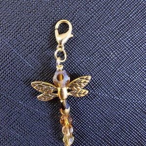 May include: A gold-toned dragonfly charm featuring a metal body with textured wings, a faceted brown bead head, and a tail composed of three graduated amber-colored crystal beads, all attached to a gold-toned lobster claw clasp for easy attachment.