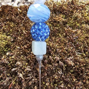 May include: A silver pin with a stack of blue and white beads. The top bead is white, the second bead is blue with white swirls, the third bead is blue with rhinestones, and the bottom bead is white.