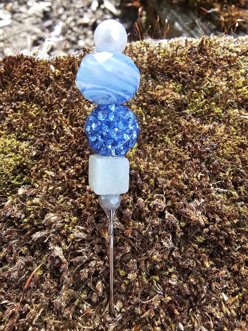 May include: A silver pin with a stack of blue and white beads. The top bead is white, the second bead is blue with white swirls, the third bead is blue with rhinestones, and the bottom bead is white.