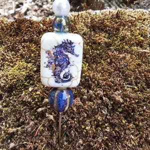 May include: A white ceramic bead with a blue and gold seahorse design, a blue and gold glass bead, and a white pearl bead on a silver pin.