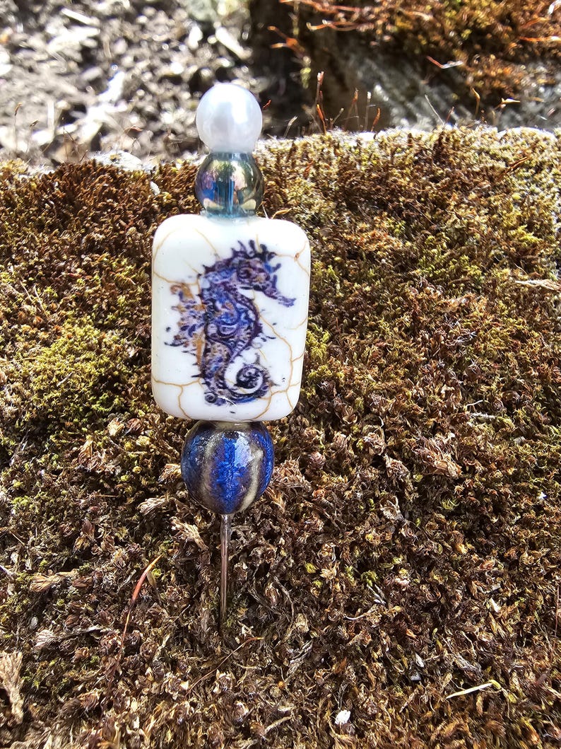 May include: A white ceramic bead with a blue and gold seahorse design, a blue and gold glass bead, and a white pearl bead on a silver pin.