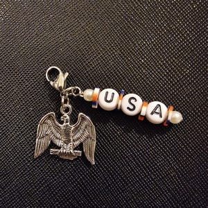 May include: A silver-colored eagle charm with outstretched wings, attached to a beaded strand spelling "USA" in white letter beads. The beads are accented with orange and blue spacers. A small pearl bead is at each end.