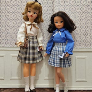 Peut inclure: Deux poupées vintage vêtues de tenues d'écolières. L'une porte une blouse blanche, une jupe à carreaux et un sac marron. L'autre porte une blouse bleue et une jupe à carreaux. Les deux poupées tiennent de petits livres et se tiennent devant un mur à motifs.