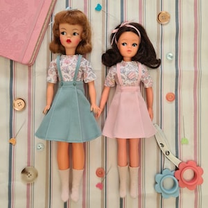 Könnte beinhalten: Zwei Vintage-Puppen in passenden Outfits mit Blusen mit Blumenmuster und einfarbigen Röcken. Die Puppe links trägt einen türkisfarbenen Rock und die Puppe rechts einen rosa Rock. Beide Puppen haben langes Haar und tragen weiße Socken.