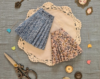 Può includere: Due gonne con stampa floreale, una blu e una rosa, sono esposte su un pizzo bianco all'uncinetto. Le gonne sono plissettate e hanno uno stile vintage.