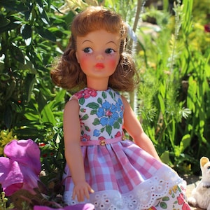 Può includere: Una bambola vintage con capelli rossi e guance rosee, indossa un vestito a fiori con una gonna a quadretti rosa. La bambola è posizionata all'aperto tra fiori e verde vibranti, con una piccola statuina di cane bianco nelle vicinanze.