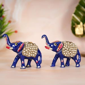May include: Two cobalt blue elephant figurines with white and gold accents. The elephants have raised trunks, red tongues, and are adorned with pearl-like embellishments on their backs. The figurines are decorative and likely for display.