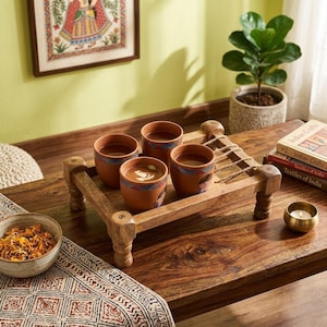 May include: A wooden serving tray holds four terracotta cups filled with a warm beverage. The cups have decorative patterns and sit on a wooden tray with a unique design. A small bowl of dried flowers and a candle are also on the table.