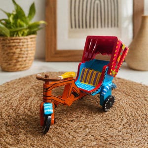 May include: A colorful, wooden rickshaw model with an orange frame, blue seat, and red canopy. The seat has yellow and purple stripes. The wheels are black, and the canopy is decorated with floral patterns. The model is displayed on a woven mat.